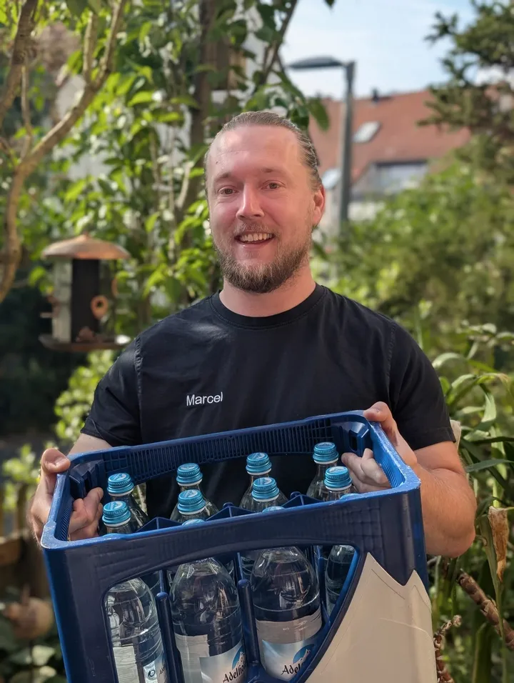 Marcel, Inhaber vom Getränkestodl, lächelt in die Kamera und trägt einen Kasten Mineralwasser